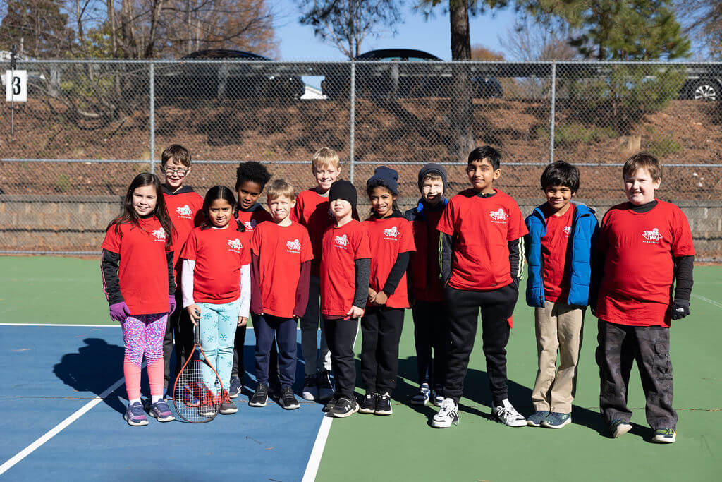 Group of smiling children wearing matching red Tennis Time Academies shirts standing together on a tennis court, posing for a group photo on a sunny day.