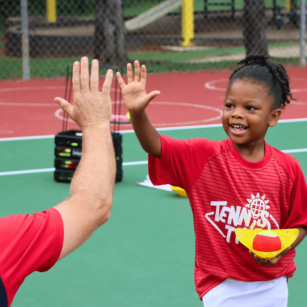 Young child smiling during a tennis class, gently high-fiving a coach after a successful rally.
