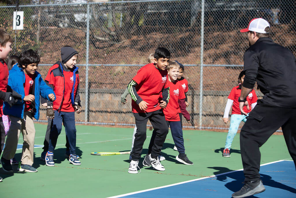 Group of young kids participating in a fun tennis warm-up drill led by a SMART Tennis™ coach at Tennis Time Academies, building coordination and teamwork on an outdoor court.