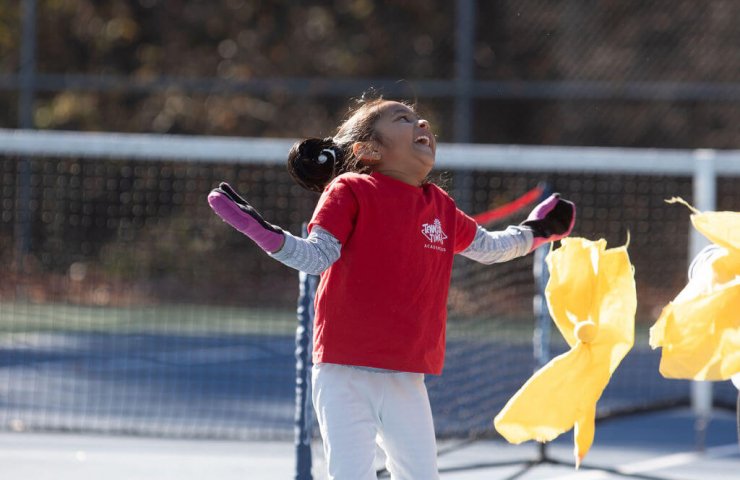 Young girl smiling mid-drill on the tennis court, celebrating a moment of joy during beginner tennis lessons for kids.