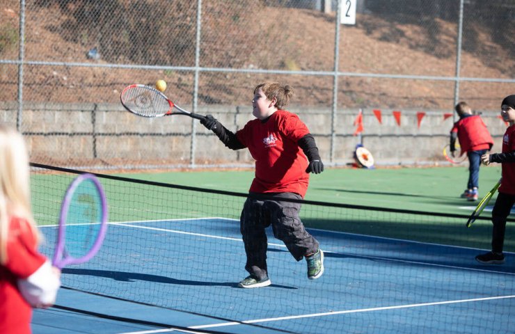 Young child swinging a foam tennis racket while balancing on one foot, smiling during a SMART Tennis™ class
