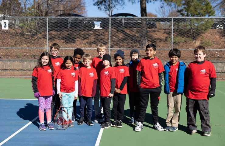 Group of smiling children wearing matching red Tennis Time Academies shirts standing together on a tennis court, posing for a group photo on a sunny day.