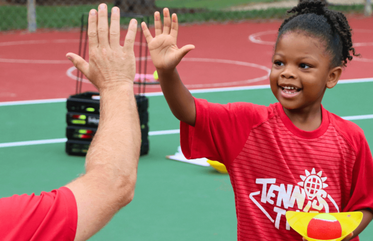 Young child smiling during a tennis class, gently high-fiving a coach after a successful rally.